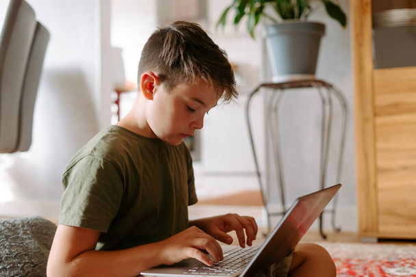 Komoly kisfiú ül a padlón, keresztbe tett lábakkal, és gépel laptop billentyűzeten otthon - Fotó, kép