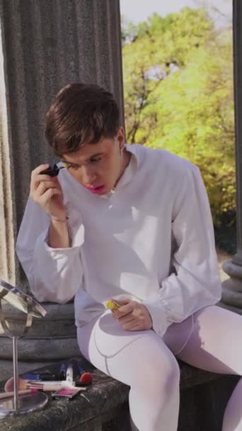 Handheld shot of young non binary person applying yellow eyeliner on eyelid and spreading powder on cheek while looking in mirror in daytime near columns - Footage, Video