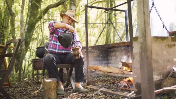 Argentyński mężczyzna wylewa gorącą wodę z czajnika do dyni kalabryjskiej przeciwko stojakowi powyżej płonącego ognia na wsi - Materiał filmowy, wideo