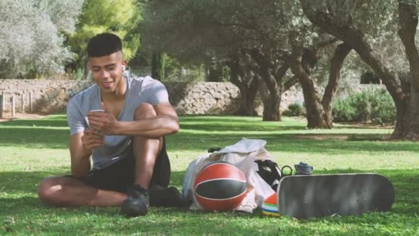 Junger ethnischer Typ in Sportbekleidung und drahtlosen Kopfhörern liest Nachrichten auf dem Smartphone und gluckst, während er auf dem Rasen in der Nähe von Ausrüstung sitzt und sich beim Training im Park ausruht - Filmmaterial, Video