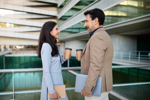 Vidám magabiztos középkorú üzletemberek elegáns férfi és nő kütyü a kezükben beszélgetnek, és ivás elvitelre kávé az utcán után sikeres üzleti találkozó - Fotó, kép