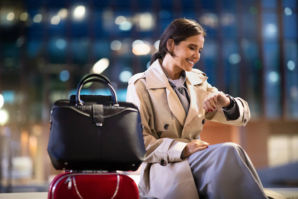 Geschäftsfrau sitzt in der Flughafenlounge und wartet auf den Flug. Glückliche Frau mittleren Alters sitzt mit Gepäck im Wartebereich des Flughafens, schaut auf die Uhr am Handgelenk und checkt die Uhrzeit. Geschäftsreisen - Foto, Bild
