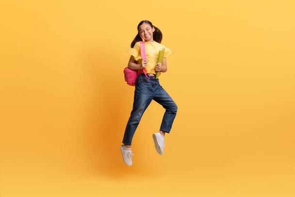 Ładna nastolatka w luźnym stroju, chodząca do szkoły, niosąca plecak, trzymająca zeszyty z ćwiczeniami, skacząca w powietrzu, odizolowana na żółtym tle, kopiująca przestrzeń. Edukacja dla dzieci, szkoła - Zdjęcie, obraz