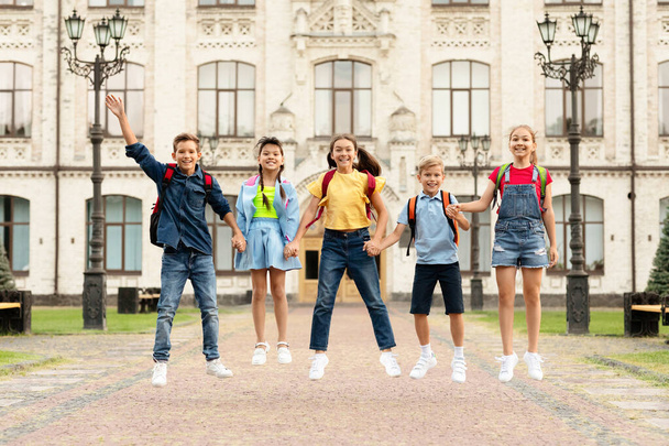 Happy Group Of Kids With Backpacks Holding Hands And Jumping Together Outdoors, Cheerful Multiethnic Boys And Girls Having Fun Against School Building, Ready For Lessons, Enjoying Education - Photo, Image