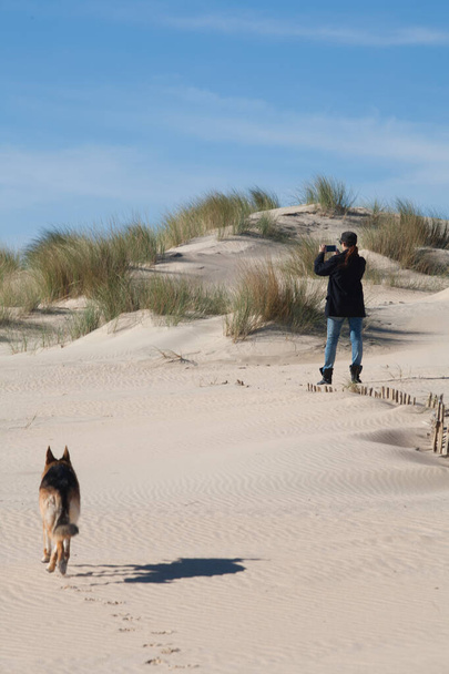 Uma mulher captura a natureza com seu smartphone em uma duna de areia, acompanhada por um cachorro. Com espaço de cópia - Foto, Imagem