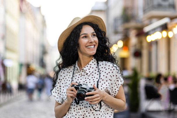 Young beautiful hispanic woman with curly hair walking in the evening city with a camera, female tourist on a trip exploring historical landmarks in the city. - Photo, Image