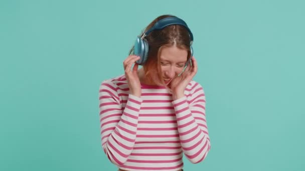 Szczęśliwa kobieta w słuchawkach słuchająca muzyki i tańcząca dyskoteka zabawiająca się gestykulacyjnymi dłońmi relaksującymi na imprezie. Ładna dziewczyna w okularach przeciwsłonecznych na niebieskim tle studio - Materiał filmowy, wideo