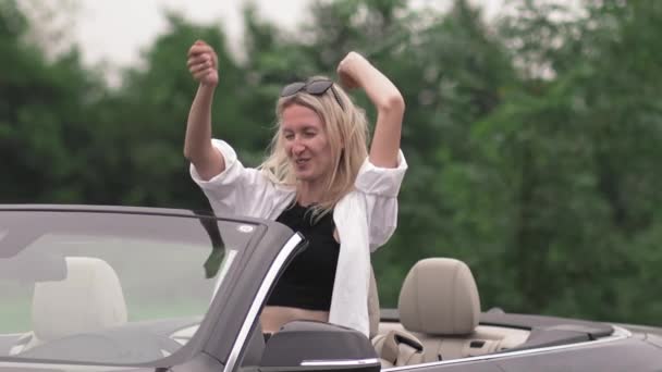 Vrouw dansend in cabrio met opgeheven handen. Handheld beelden van jonge vrouw genieten van winderige dag op reis in de zomer. Ze heeft plezier met reizen.. - Video