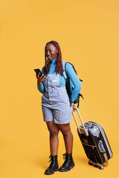 African american girl texting messages and carrying trolley bags in studio, having fun on vacation journey. Female traveller browsing online app on mobile phone website, holiday trip. - Photo, Image