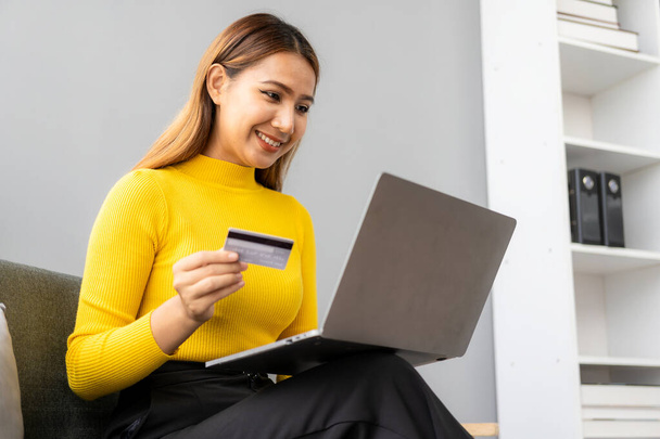 Asiatisches Mädchen bezahlt online per Laptop, um zu Hause einzukaufen Attraktive asiatische Millennial-Frau hält Smartphone und Kreditkarte per Mobile Banking oder Online-Shopping-App in der Hand. - Foto, Bild