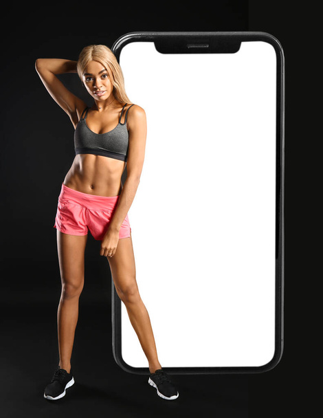Sportieve Afro-Amerikaanse vrouw en grote smartphone op donkere achtergrond. Gewichtsverlies concept - Foto, afbeelding