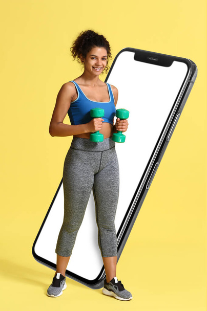 Спортивная афроамериканка с гантелями и большим смартфоном на жёлтом фоне. Концепция потери веса - Фото, изображение