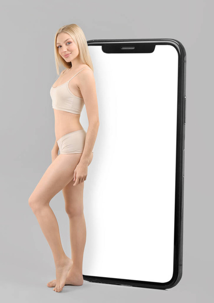 Schlanke Frau in Unterwäsche und großem Smartphone auf grauem Hintergrund. Konzept zur Gewichtsreduktion - Foto, Bild