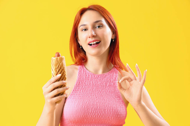 Όμορφη νεαρή γυναίκα με νόστιμο γαλλικό χοτ ντογκ δείχνει OK σε κίτρινο φόντο - Φωτογραφία, εικόνα