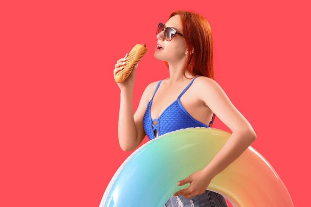 Beautiful young woman with french hot dog and inflatable ring on red background - Photo, Image