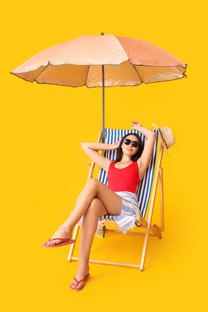 Schöne junge Frau mit Regenschirm sitzt auf Liegestuhl vor gelbem Hintergrund - Foto, Bild