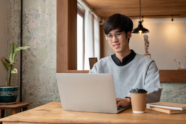 A happy and smart young Asian male college student in casual clothes and eyeglasses is using his laptop computer while sitting in a coffee shop. - Photo, Image