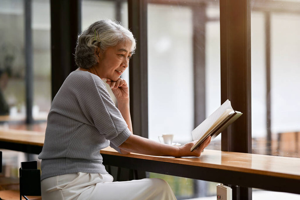 Een gelukkige en ontspannen Aziatische gepensioneerde dame leest een boek terwijl ze aan een tafel zit in een koffieshop en haar weekend alleen door brengt in de stad. Pensioenleefstijlconcept - Foto, afbeelding