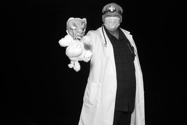 Een kwade dokter poseert met griezelig speelgoed in zijn hand als hij zijn foto genomen terwijl in een Halloween Photo Booth met zwart fluwelen gordijnen. - Foto, afbeelding