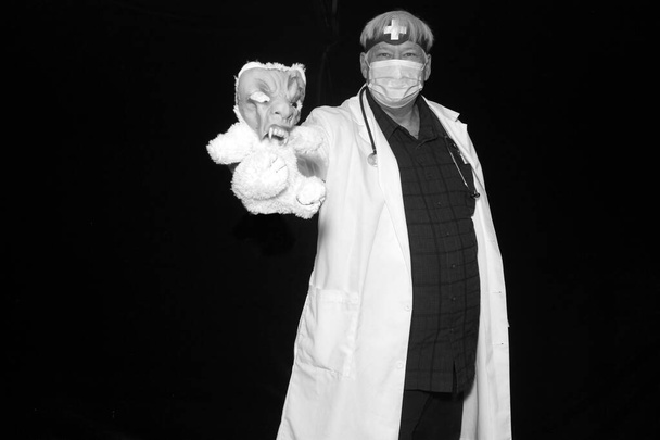 Een kwade dokter poseert met griezelig speelgoed in zijn hand als hij zijn foto genomen terwijl in een Halloween Photo Booth met zwart fluwelen gordijnen. - Foto, afbeelding