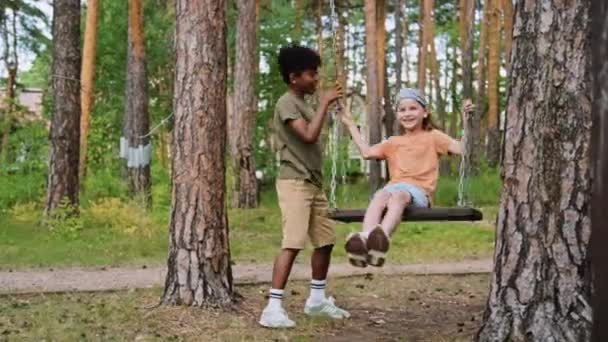 Full tracking shot of African American and Caucasian elementary age boys playing on swing in forest on playground - Footage, Video