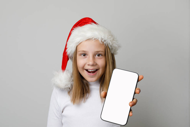 Santa dziewczyna pokazuje smartfon z pustym białym ekranem wyświetlacza. Koncepcja świąteczna - Zdjęcie, obraz