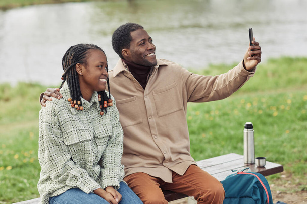 Ευτυχισμένο ζευγάρι Αφρικανών χαμογελά στην κάμερα κάνοντας selfie πορτρέτο στο κινητό τηλέφωνο, ενώ κάθεται στον πάγκο στο πάρκο - Φωτογραφία, εικόνα