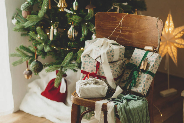 Wesołych Świąt i Wesołych Świąt! Stylowe pakowane prezenty świąteczne z wstążką na drewnianym starym krześle na tle ozdobionej choinki z vintage bombki i świąteczne światła - Zdjęcie, obraz