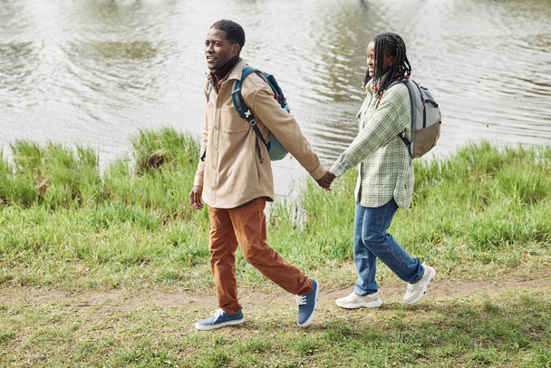 African teenage couple with backpacks holding hands while walking along the bank of the river during their walking - Photo, Image