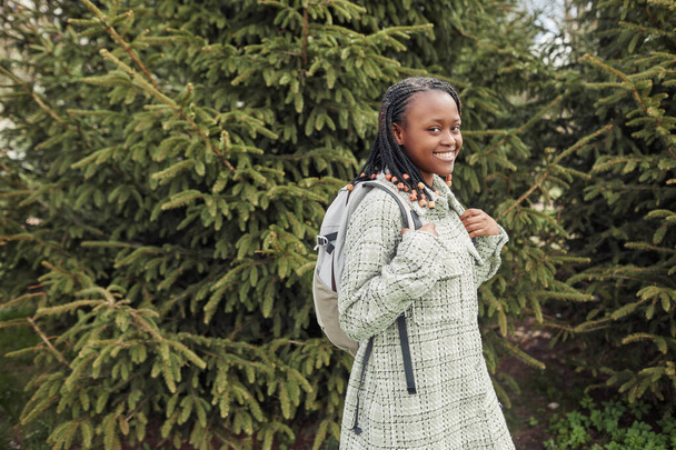 Dışarıda çam ağaçlarının arasında yürürken sırt çantasıyla kameraya gülümseyen Afrikalı genç kızın portresi. - Fotoğraf, Görsel