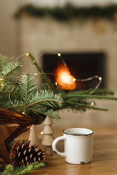 Stilvolle Tasse warmen Tee, Tannenzweige, Holzbäume und Sterne, Tannenzapfen auf dem Tisch gegen den brennenden Kamin. Modernes weihnachtliches rustikales, umweltfreundliches Dekor und leerer Becher, skandinavische Winterhygge - Foto, Bild