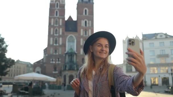 Bella donna caucasica elegante in un cappello luminoso che tiene il telefono cellulare e scattare selfie foto o videochiamate sulla piazza del mercato della città vecchia di Cracovia  - Filmati, video