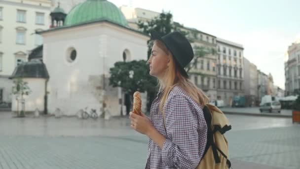 Ευτυχισμένη νεαρή ξανθιά τουρίστρια με κομψά ρούχα και ένα καπέλο Boho τρώει bagel obwarzanek παραδοσιακή πολωνική κουζίνα σνακ waling στην πλατεία Αγοράς στην Κρακοβία. Ταξιδεύοντας στην Ευρώπη. Βασιλική της Αγίας Μαρίας - Πλάνα, βίντεο