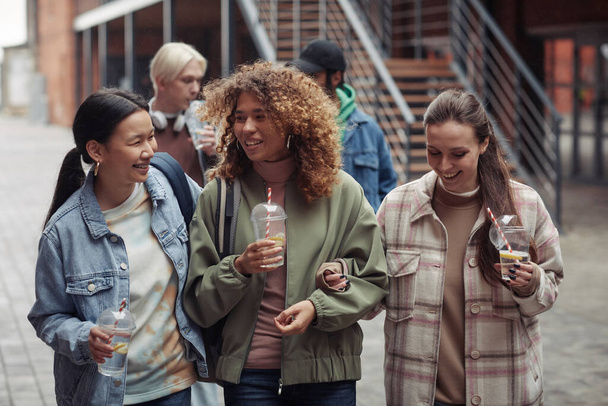 Gruppe fröhlicher interkultureller Teenager-Mädchen mit Limo, die sich unterhalten, während sie vor ihren Freunden im Freien die Straße hinunterlaufen - Foto, Bild