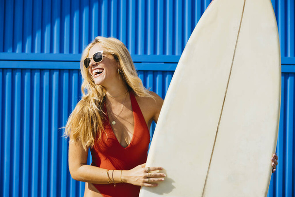 Bir sörfçünün portresi ayakta, mayolu mavi bir arka planda, yanında sörf tahtasıyla. Yaz tatilinde plaj aktiviteleri yapan mutlu bir kadın.. - Fotoğraf, Görsel