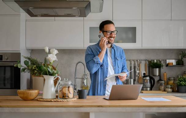 Empresário sorridente discutindo sobre telefone inteligente enquanto trabalhava no laptop no balcão da cozinha em casa - Foto, Imagem