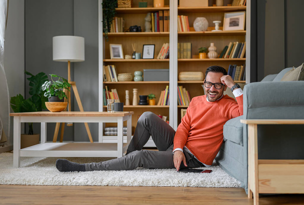 Longitud completa de joven empresario feliz con tableta digital sentado en la alfombra por el sofá en la sala de estar - Foto, imagen