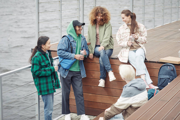 Пять веселых друзей-подростков в повседневной одежде рассказывают шутки или обсуждают смешные вещи во время отдыха на деревянном пирсе у воды после колледжа - Фото, изображение