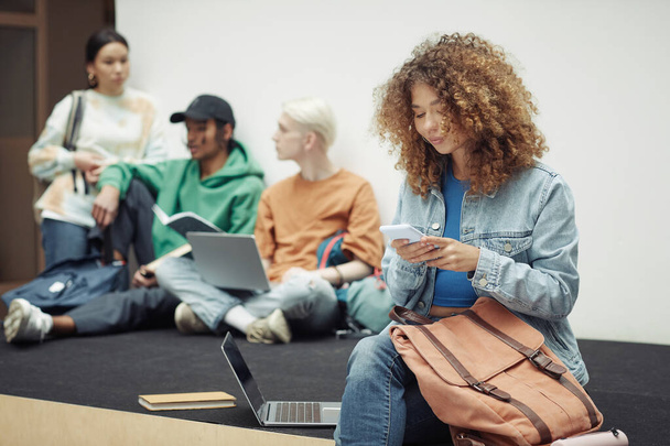 Hübsches Teenager-Mädchen in Freizeitkleidung textet in der Pause im Smartphone, während sie im Hörsaal gegen ihre Klassenkameraden sitzt und über Vorlesungsnotizen diskutiert - Foto, Bild