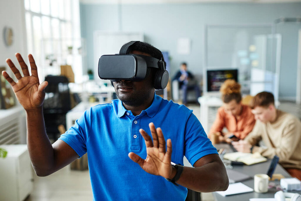 Африканский молодой человек в очках виртуальной реальности жестикулирует во время тестирования программы виртуальной реальности в офисе во время встречи с коллегами - Фото, изображение