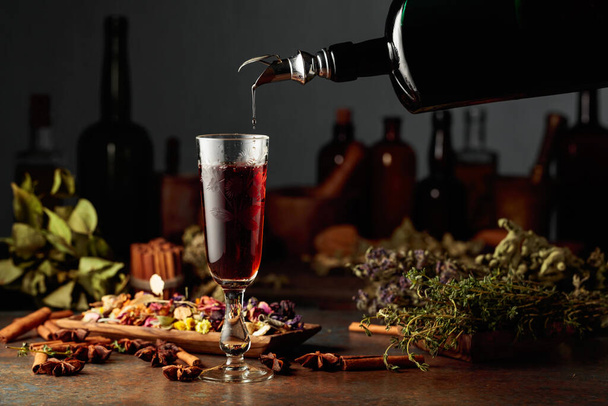 La liqueur ou le baume brun à base de plantes est versé d'une bouteille vintage dans un verre. Sur une table des herbes séchées, des fleurs, des épices et de vieux ustensiles de cuisine. Concept de phytothérapie. - Photo, image
