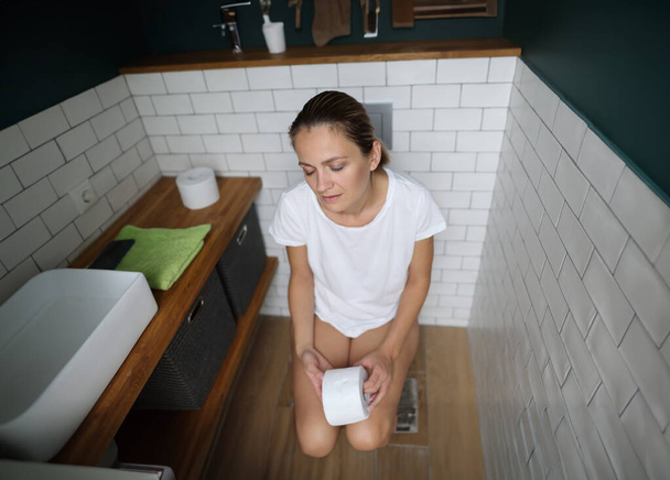 Zmęczona kobieta siedzi na toalecie i trzyma papier toaletowy. Koncepcja zaburzeń jelit - Zdjęcie, obraz