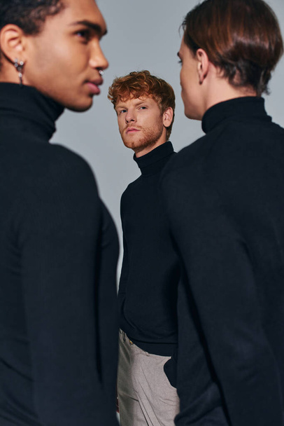 enfoque en el hombre joven de pie cerca borrosa amigos interracial sobre fondo gris, moda, vertical - Foto, Imagen
