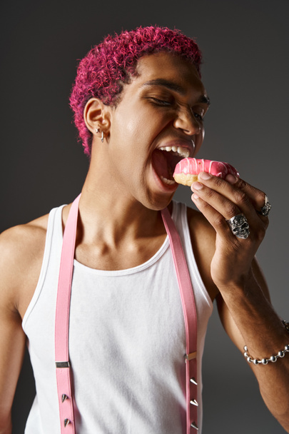 portret młodego różowłosego mężczyzny z różowymi szelkami jedzącego różowe smaczne pączki, modę i styl - Zdjęcie, obraz