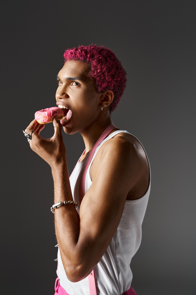 όμορφο ανδρικό μοντέλο με ροζ μαλλιά τρώγοντας ντόνατ ποζάροντας σε γκρι φόντο, μόδα και στυλ - Φωτογραφία, εικόνα