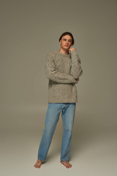 aantrekkelijke elegante man in stijlvolle gezellige trui en jeans poseren met de hand in de buurt gezicht en weg te kijken - Foto, afbeelding