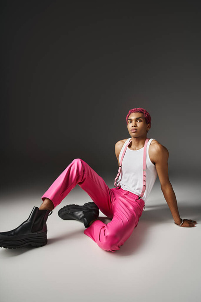 hombre afroamericano bien parecido sentado en el suelo en pantalones de color rosa con tirantes, concepto de moda - Foto, imagen