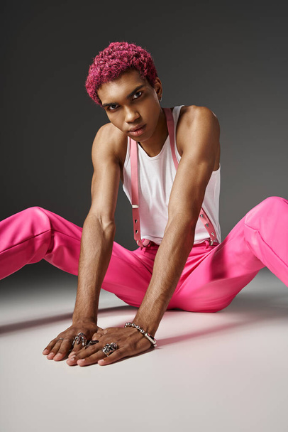 pink haired african american man in vibrant attire sitting on floor and looking at camera, fashion - Photo, Image