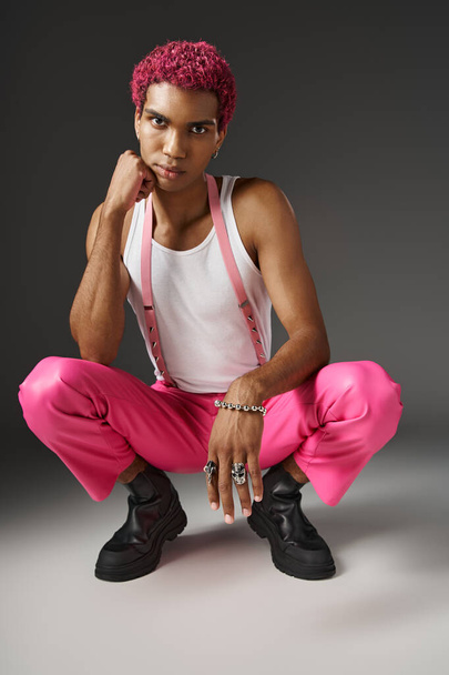 trendy african american man posing on gray backdrop squatting and looking at camera, fashion concept - Photo, Image