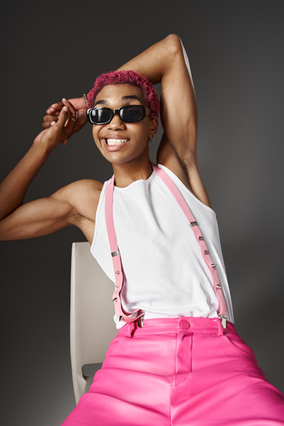 verspielter junger Mann mit rosa Haaren und Hosenträgern, der mit Sonnenbrille lächelt, Modekonzept - Foto, Bild